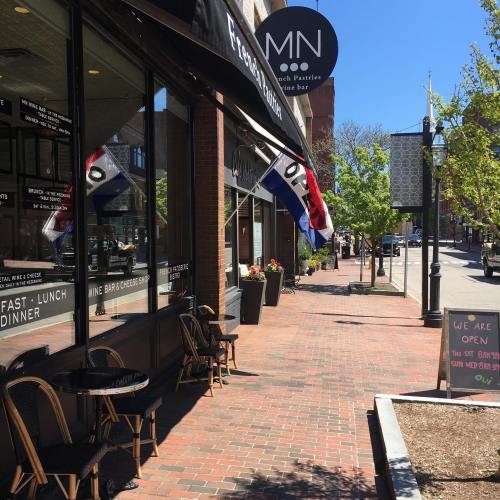 A street view of a café with outdoor seating, flags, and a sign indicating it's open on a sunny day.
