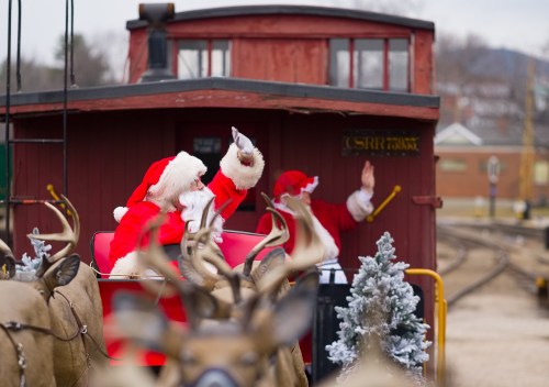 Two Santa Clauses wave from a festive train car, surrounded by reindeer and holiday decorations.