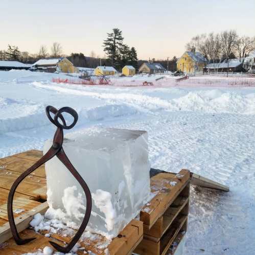 A block of ice on wooden pallets with ice tongs, set against a snowy landscape and distant yellow buildings.