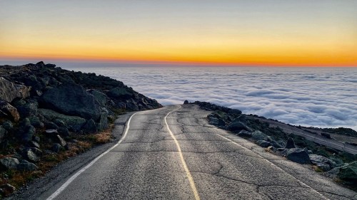 A winding road leads through rocky terrain above a sea of clouds, with a colorful sunset in the background.