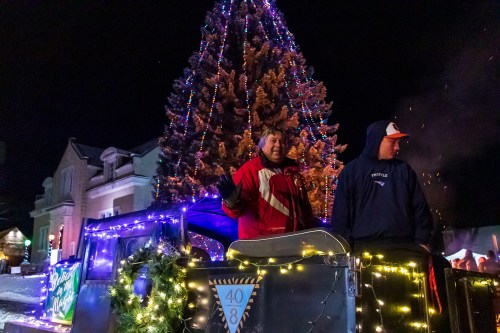 Two people on a decorated float with a large, lit Christmas tree in the background during a nighttime event.
