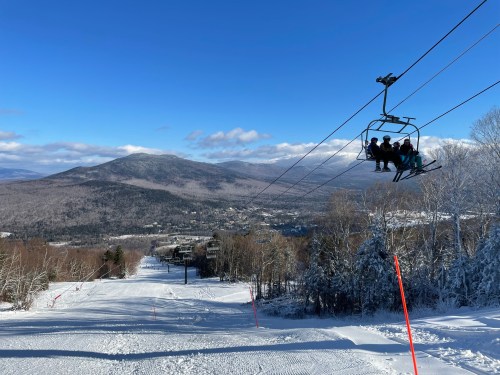 A ski lift carries passengers over a snowy landscape with mountains and a clear blue sky in the background.