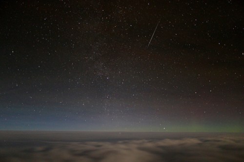 A starry night sky above a layer of clouds, with a faint aurora and a shooting star visible.
