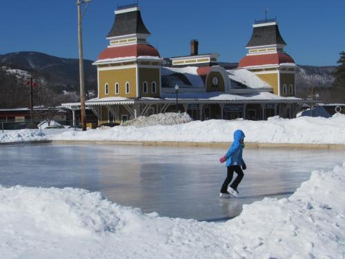 A person in a blue jacket skates on an ice rink surrounded by snow, with a historic building in the background.