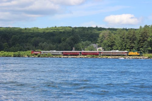 A red and yellow train travels along a riverbank, surrounded by lush green trees under a blue sky.