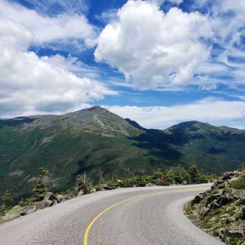 A winding road curves through a mountainous landscape under a blue sky with fluffy clouds.