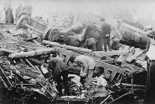 Workers and onlookers inspect the wreckage of a train derailment, with overturned cars and debris scattered around.