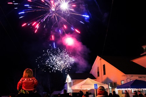 A child on an adult's shoulders watches colorful fireworks illuminate the night sky above a festive gathering.