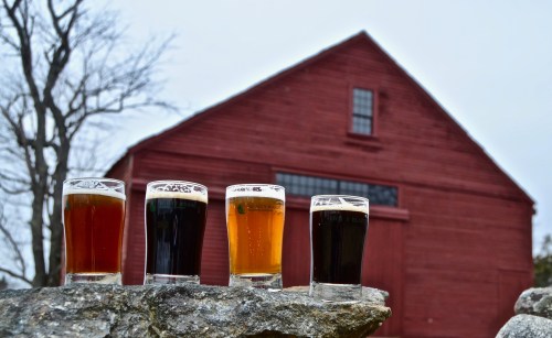 Four glasses of beer in varying colors sit on a stone ledge, with a red barn in the background.