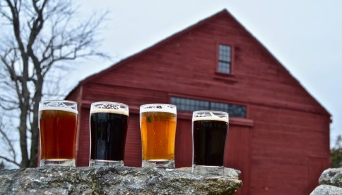 Four glasses of beer in varying colors sit on a stone ledge, with a red barn in the background.