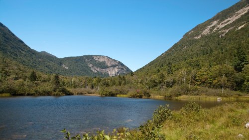 A serene landscape featuring a calm lake surrounded by lush greenery and mountains under a clear blue sky.