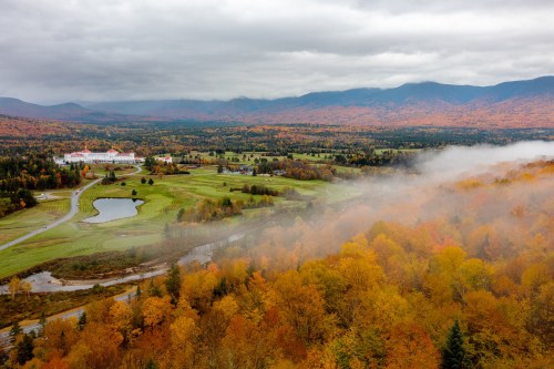 Aerial view of a colorful autumn landscape with mountains, a golf course, and mist over trees in vibrant fall foliage.