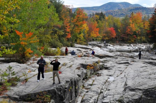 People exploring a rocky landscape surrounded by vibrant autumn foliage and mountains in the background.
