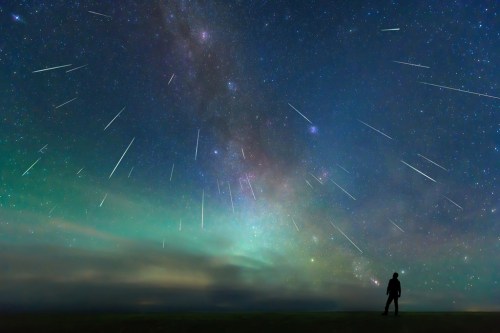 A silhouette of a person gazes up at a starry sky filled with shooting stars and vibrant colors.