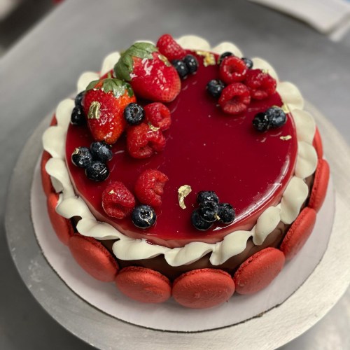 A round cake topped with strawberries, raspberries, blueberries, and a glossy red glaze, surrounded by red macarons.