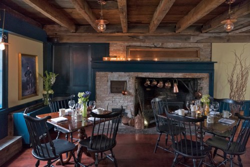 Cozy restaurant interior with wooden beams, a fireplace, and tables set for dining, adorned with flowers and candles.