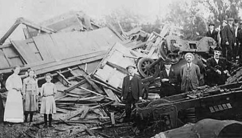 Historical photo of a group of people standing near a collapsed building and wreckage, with trees in the background.