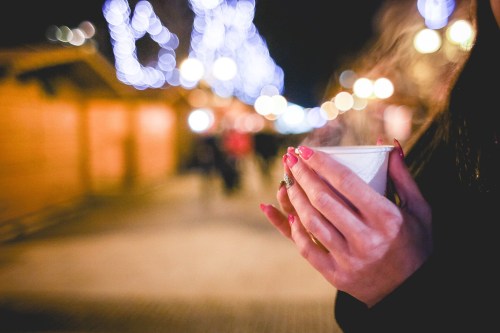 A person holds a steaming cup in a festive, softly lit outdoor market with blurred lights in the background.