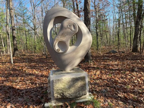 A modern sculpture on a stone pedestal, surrounded by trees and fallen leaves in a wooded area.