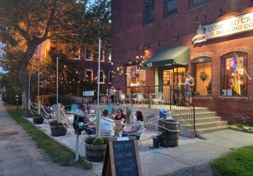A cozy brewery patio with string lights, people enjoying drinks, and a welcoming entrance on a warm evening.
