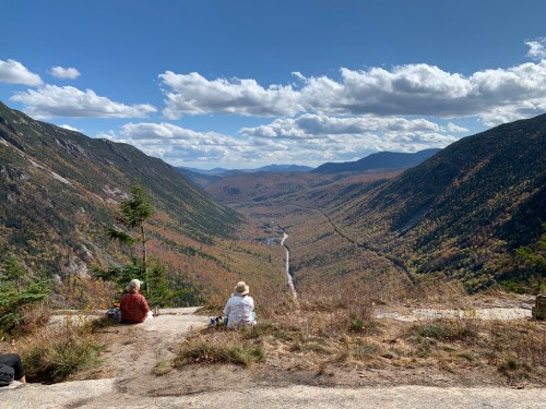 Two people sit on a rocky ledge, overlooking a valley filled with autumn foliage and mountains under a blue sky.