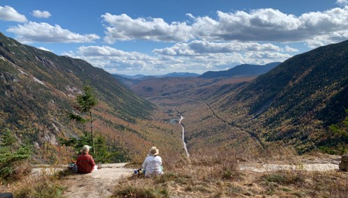 Two people sit on a rocky ledge, overlooking a colorful valley with mountains and a winding river under a blue sky.