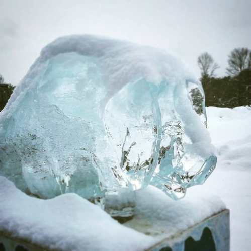 A clear ice sculpture of a bear, partially covered in snow, set against a winter landscape.