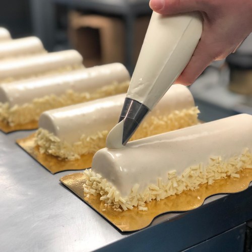 A pastry chef decorates a white mousse cake with a piping bag, surrounded by chocolate shavings on a gold base.