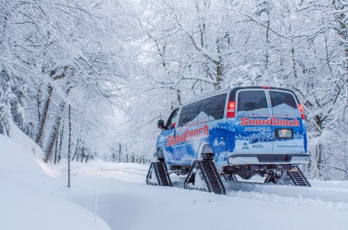 A blue snow coach vehicle driving through a snowy, tree-lined path in a winter landscape.