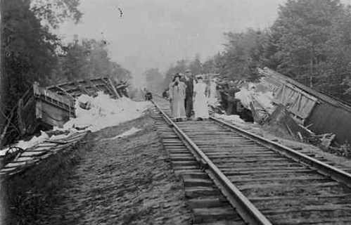 Historic black-and-white photo of a train derailment, with damaged cars off the tracks and people inspecting the scene.