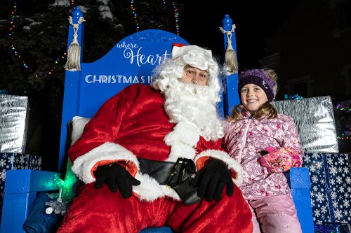 A smiling child in winter attire sits next to Santa Claus on a decorated chair, surrounded by festive lights and gifts.