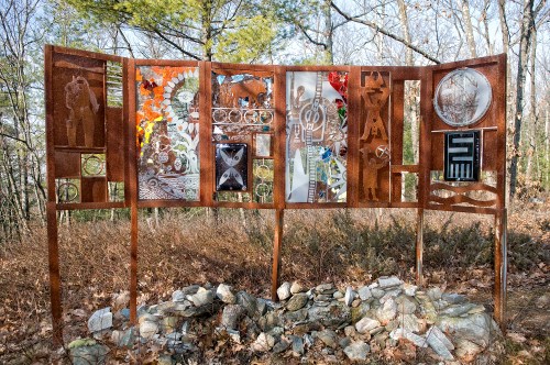 A rusted metal art installation featuring various panels with intricate designs, set in a wooded area.