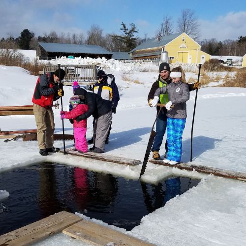 A group of five people in winter clothing stand by a frozen lake, preparing to ice fish. Snowy landscape in the background.