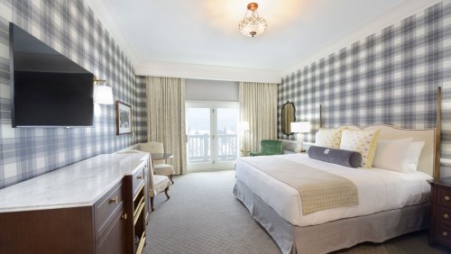 A stylish hotel room featuring a king bed, plaid wallpaper, a TV, and a balcony with scenic views.