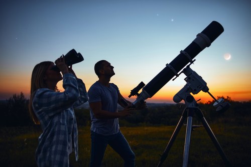 A man uses a telescope while a woman observes the sky with binoculars at sunset. Stars and planets are visible.