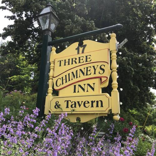 Sign for "Three Chimneys Inn & Tavern" with a yellow background, surrounded by flowers and greenery.