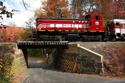 A red train engine passes over a wooden bridge, surrounded by autumn foliage and a quiet road below.