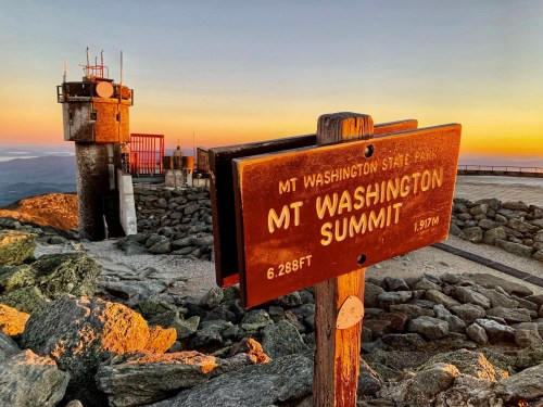 Sign at Mt. Washington Summit with rocky terrain and a tower in the background, illuminated by a colorful sunset.