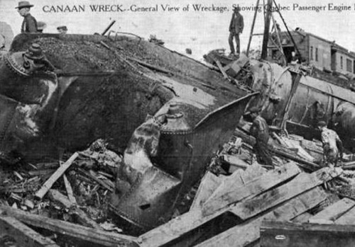 Black and white image of a train wreck, with people inspecting the damaged locomotive and debris scattered around.