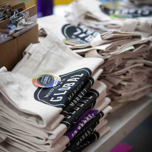 A stack of reusable tote bags with a "Global Market" logo and colorful buttons on a table.