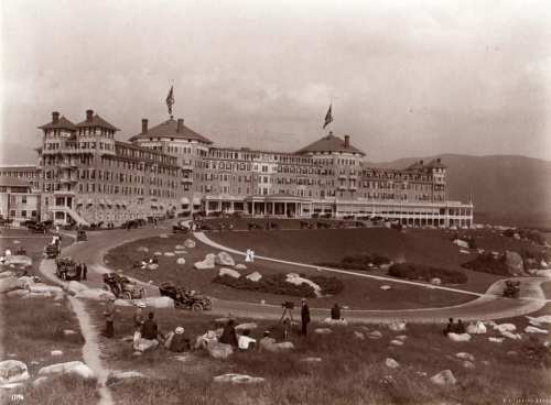 Historic black-and-white photo of a large hotel with flags, surrounded by cars and people on a grassy area.
