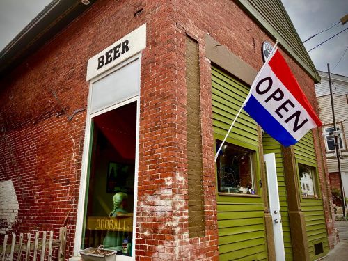 A brick building with a "Beer" sign and an "Open" flag, featuring a green door and a window displaying a statue.