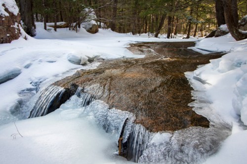 A serene winter scene featuring a flowing stream surrounded by snow and ice, with trees in the background.