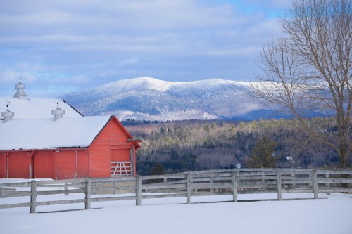 A red barn in a snowy landscape with mountains in the background under a partly cloudy sky.