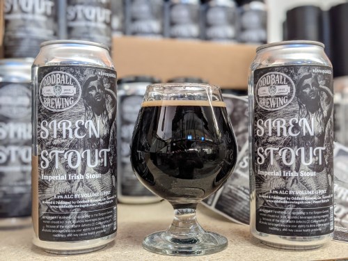 Two cans of Siren Stout beside a glass of dark stout, with a backdrop of more cans in a brewery setting.