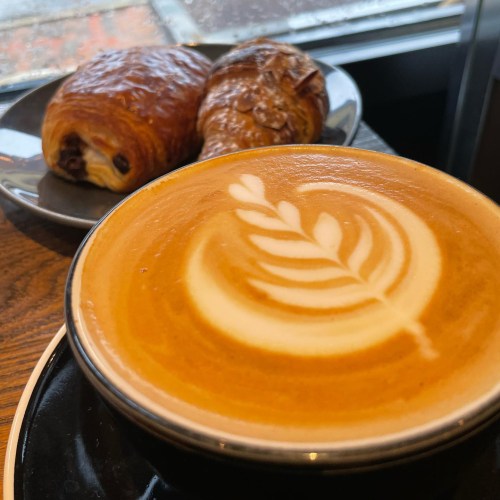 A latte with a leaf design in foam, accompanied by a chocolate croissant and a pastry on a plate.