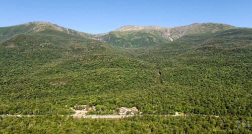 Aerial view of lush green mountains with a small building nestled at the base, under a clear blue sky.