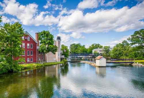 A serene river scene with a red building, lush trees, and a small white structure on the water under a blue sky.