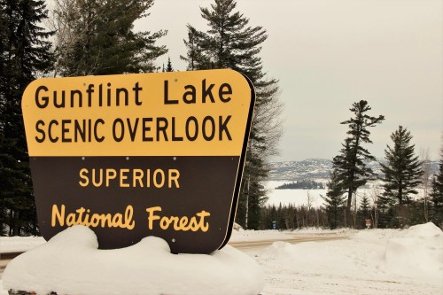 Sign for Gunflint Lake Scenic Overlook in winter, surrounded by snow and trees, with a view of the lake in the background.