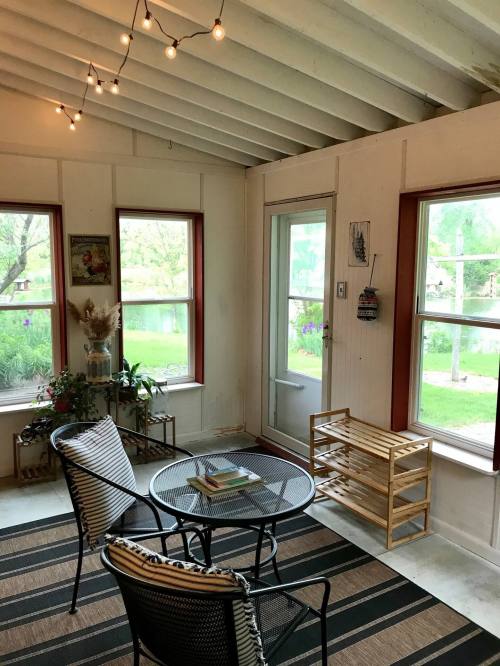 Cozy sunroom with string lights, a round table, two chairs, and large windows overlooking a garden.